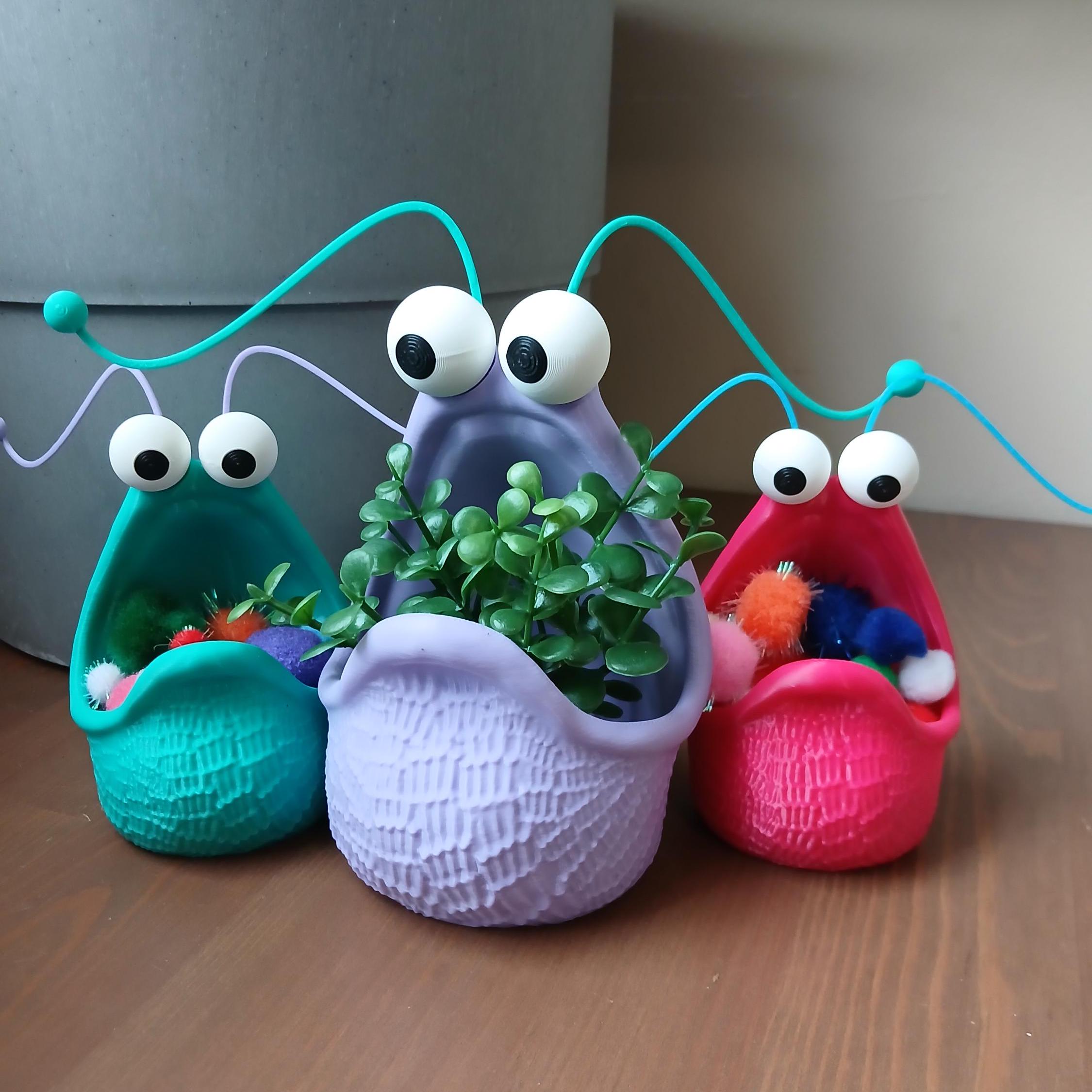 A set of 3 planters, in cyan, purple, and pink. Two outside ones are holding pom poms and the center one is holding a succulent.