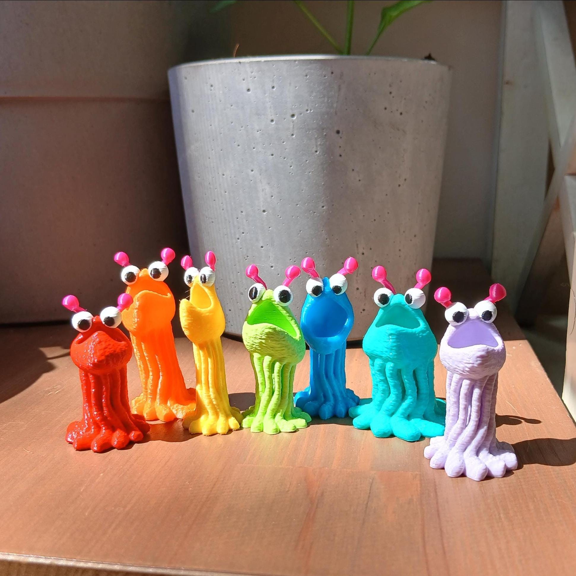Colorful toy monsters on a wooden surface with a plant pot in the background. Seven in total, each a different color of the rainbow