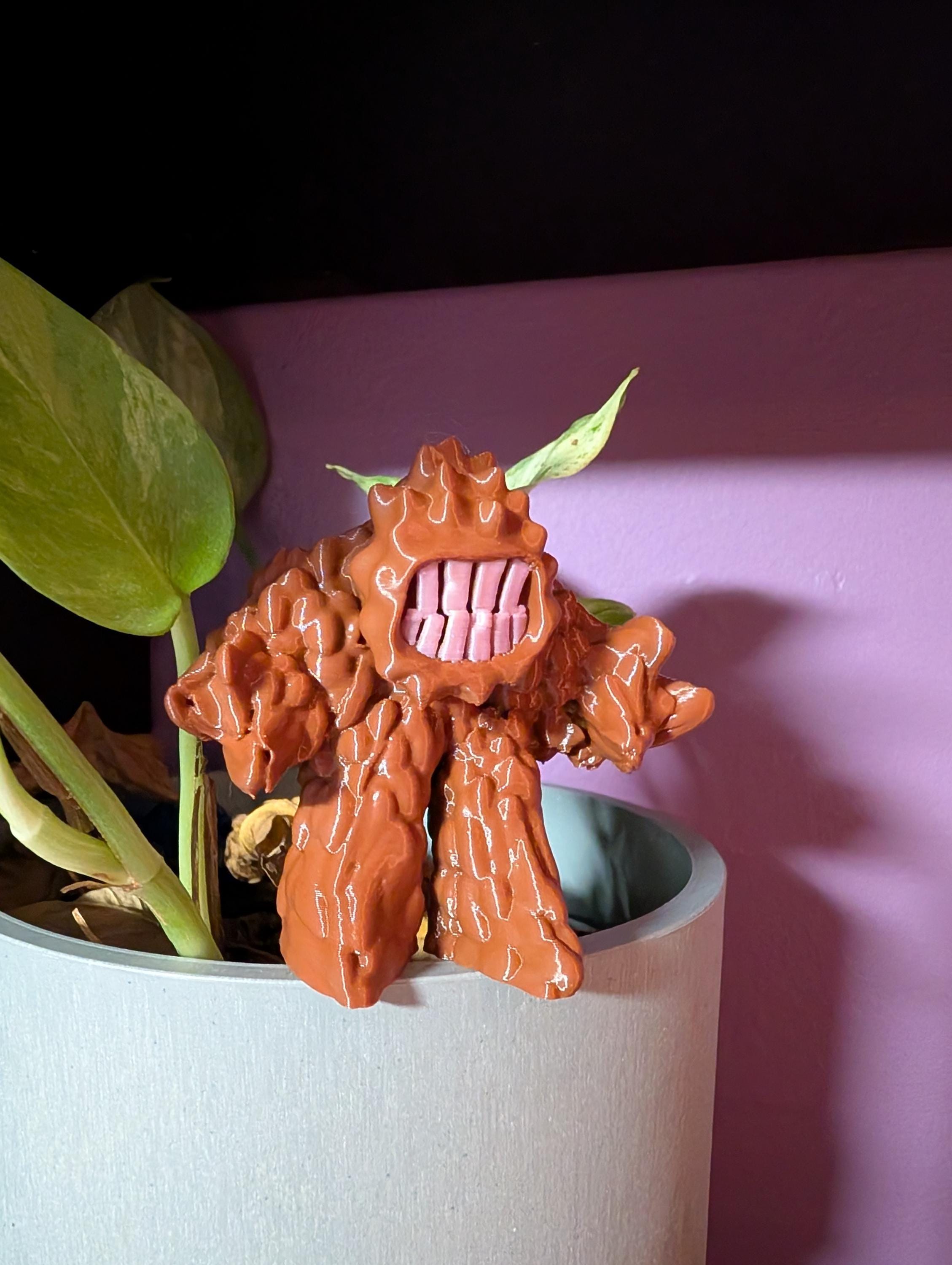 3D printed Mud Monster figurine with big teeth and bumpy body, cute but mischievous desk or shelf decor.  Shown balancing on the edge of a planter.