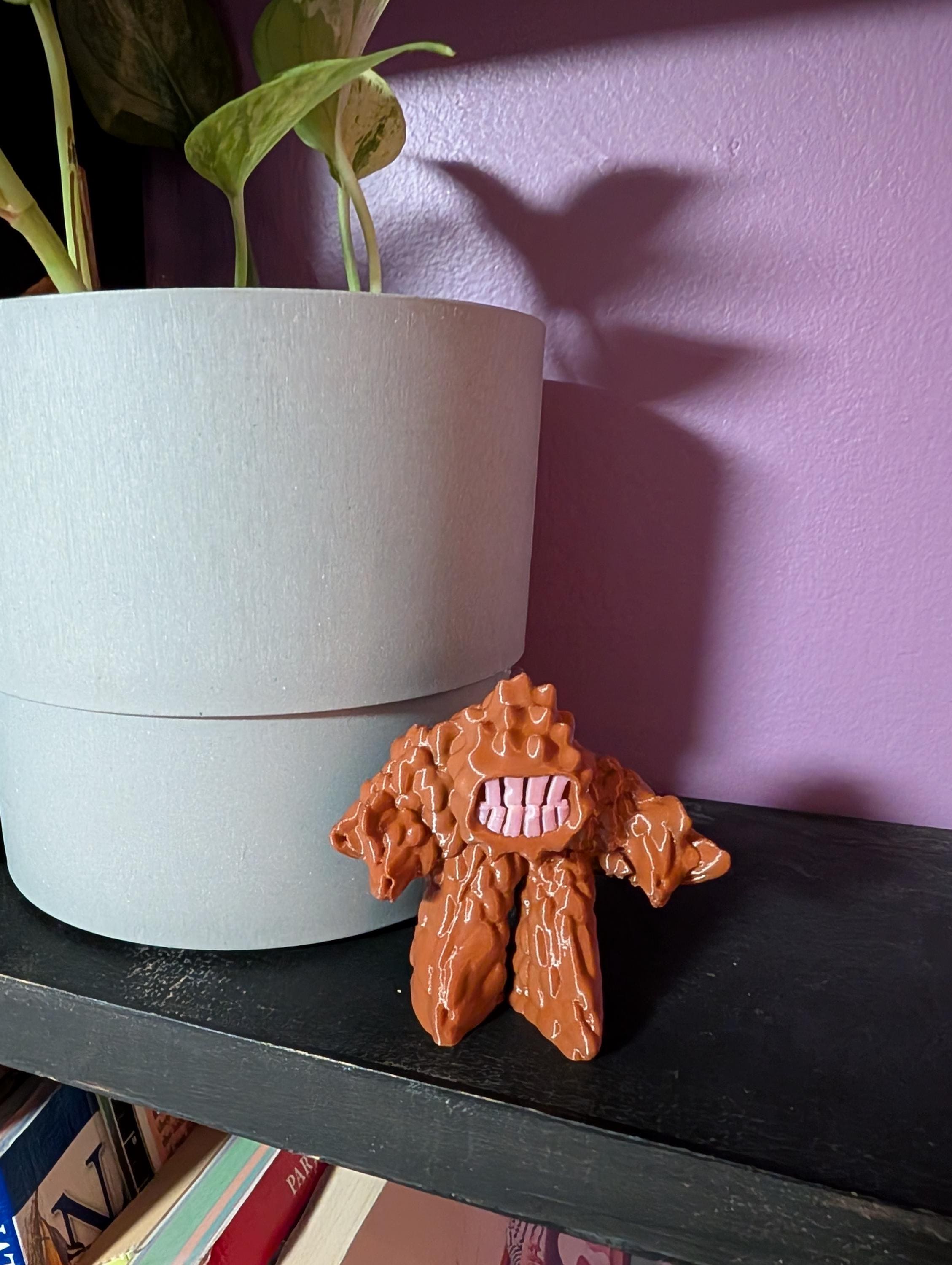 3D printed Mud Monster figurine with big teeth and bumpy body, cute but mischievous desk or shelf decor. Shown beside a planter