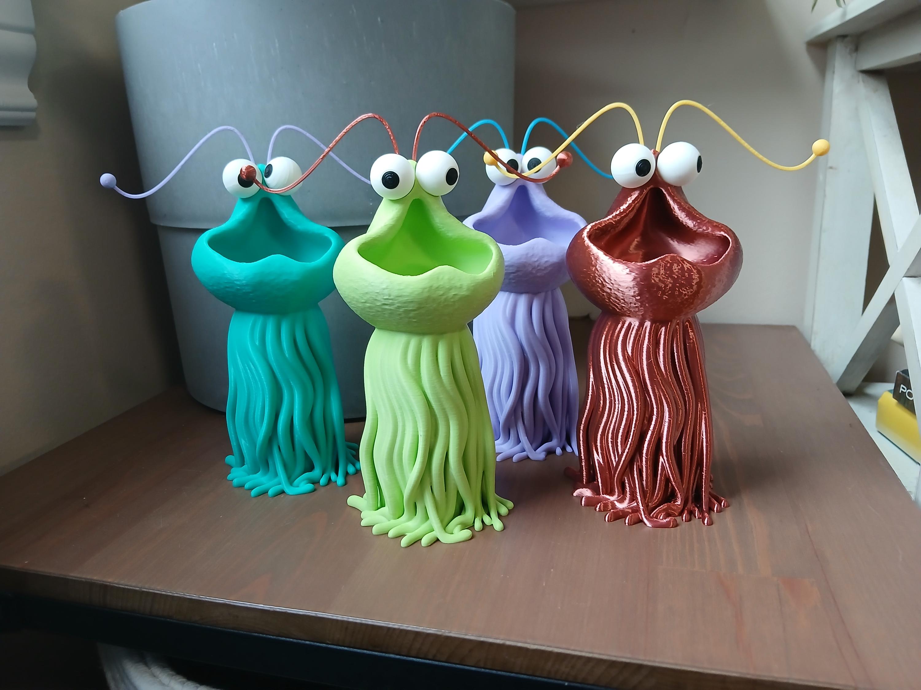Four colorful alien-like vase figurines on a wooden surface with a neutral background. Vases have detachable antennas and big round eyes that can be adjusted.